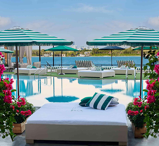 Daybed by pool with green and white striped pillows, surrounded by pink flowers, with Biscayne Bay in the background