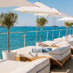 Outdoor patio with lounge chairs lined up with white umbrellas overhead and a blue sea in the background