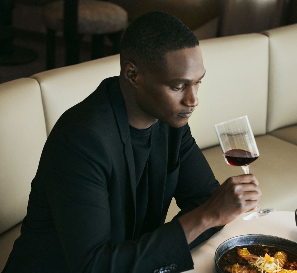 Polished looking man in a black suit sits at a dining table, holding a glass of red wine