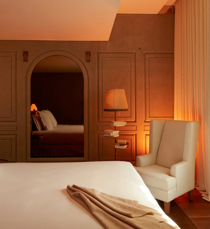 A freshly made white bed in the foreground, captured in an arched mirror against the far wall, where there is also an armchair and floor lamp