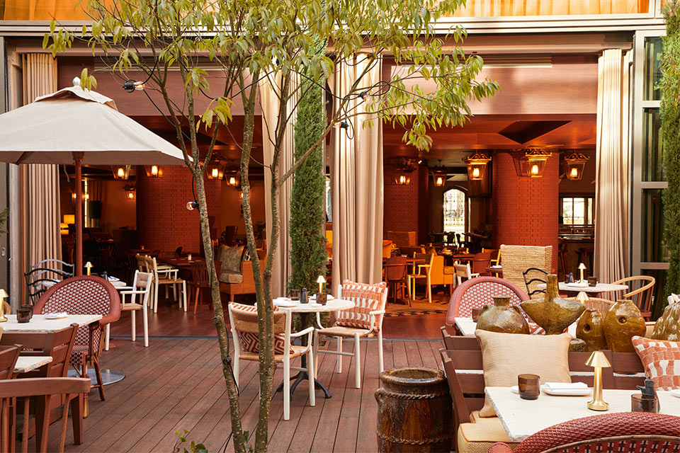 Beautiful outdoor terrace with small tables, cushioned seats, umbrellas, greenery, and open doors looking into a large dining space. 