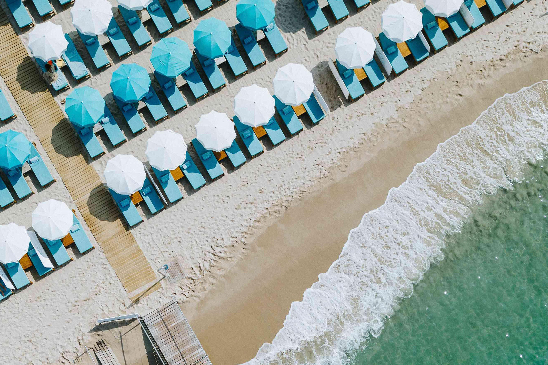 Aerial view of a sandy beach with turquoise waters and teal chaise lounges with white and teal umbrellas