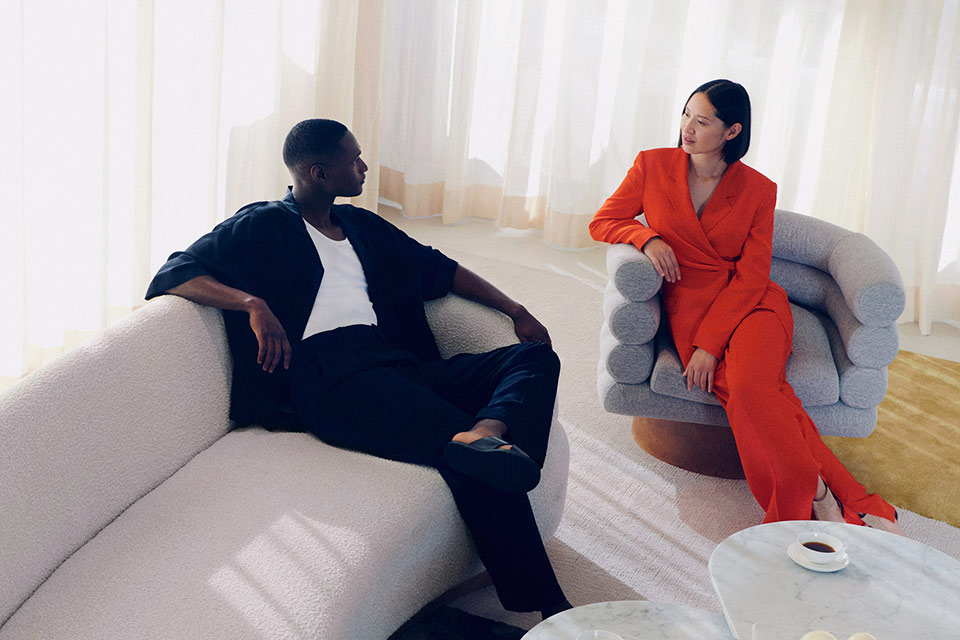 A man dressed in a black two piece outfit, and a woman in an orange jumpsuit, sitting in chairs enjoying a cup of coffee.