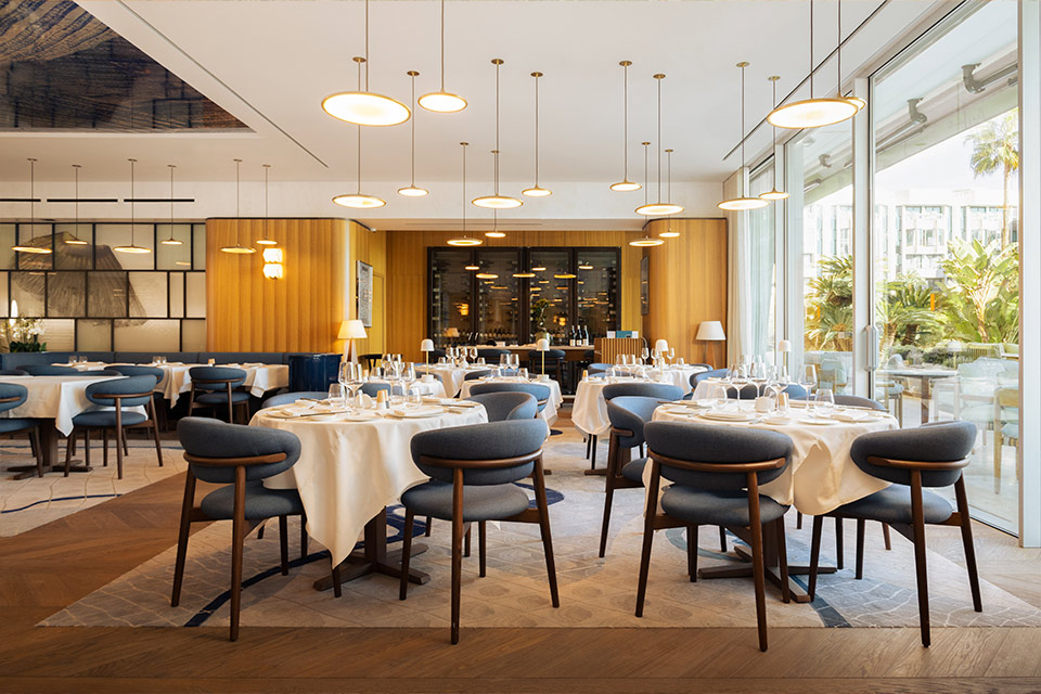 Large restaurant dining room with small cream-colored tablecloth tables with dark blue and wooden dining chairs, pendant lights hanging overhead, with a wooden floor with a large rug.