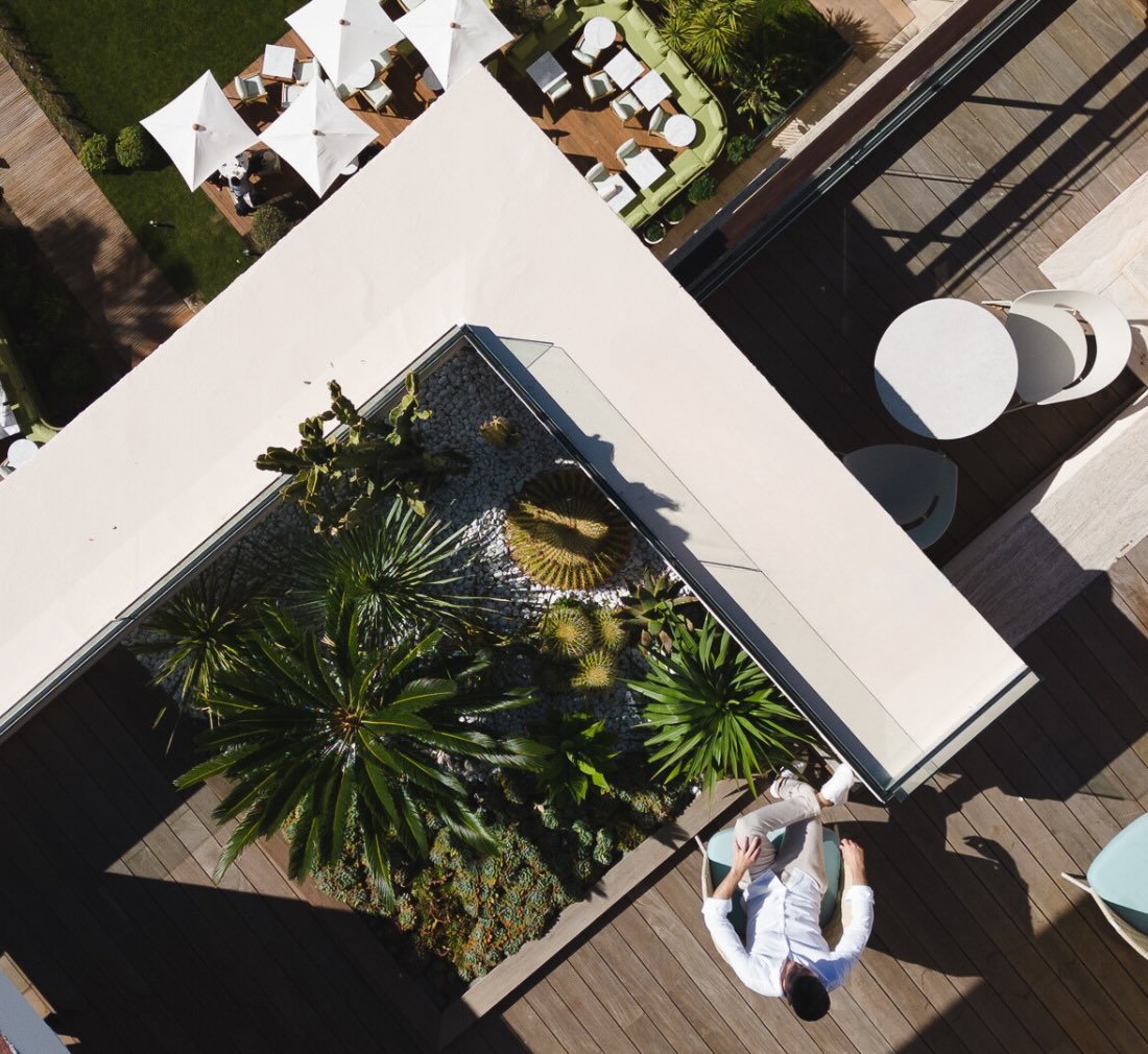 An elevated terrace captured from above in the daytime, showing a man sitting in a chair alongside a small garden of palms. On the ground in the distance is a patio with tables and umbrellas.