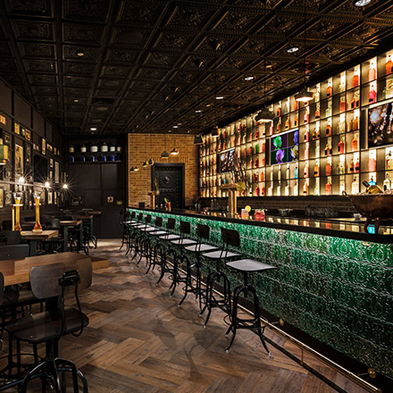 A long, dimly lit bar with black high chairs, a fully stocked bar and black tin ceiling at Hudson Tavern Doha.