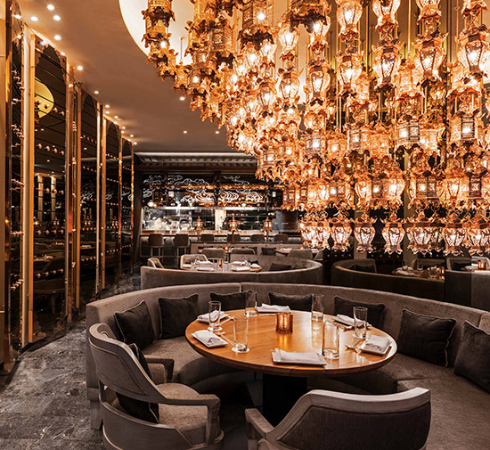 Elegant dining room with a large intricate golden chandelier over a round wooden table with grey banquette seating and two grey dining chairs. 