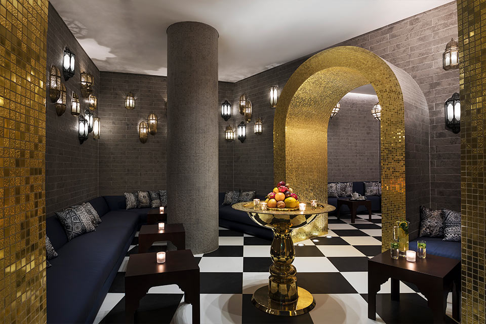 Mondrian Doha spa with black and white checkered floor, gold and black tiled walls, a blue wraparound couch, and a gold tray in the middle of the room with fresh fruit.