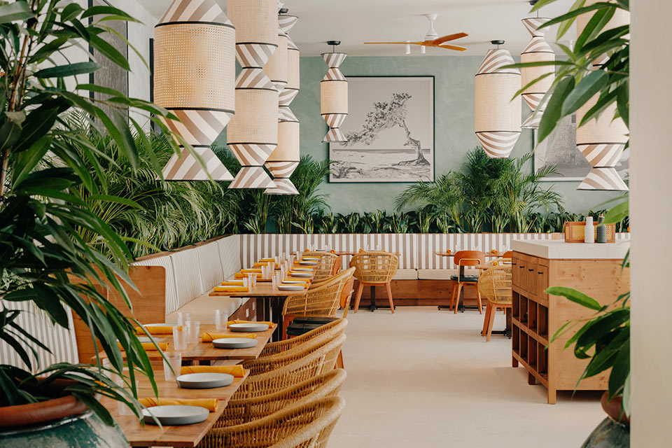 Dining space with square wooden tables, wicker chairs, green plants scattered throughout, and hanging pendant lights with striped design. 