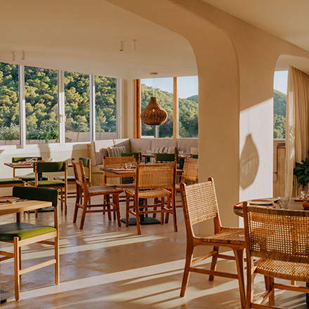 Bright dining space with natural light, wicker dining chairs, green dining chairs, and wooden tables with windows looking out to a green forest.