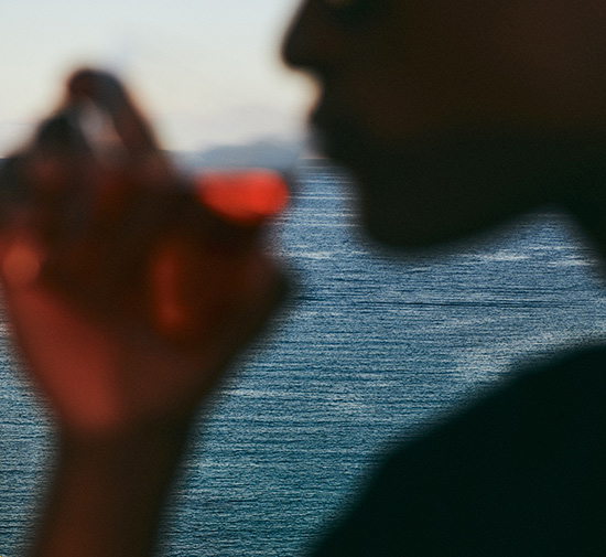 Close-up blurred image of man sipping a red cocktail with a blue ocean in the background.