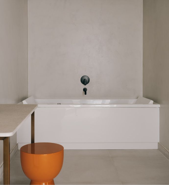 Large whigte bathtub in a cream colored bathroom with an orange stool and modern vanity in the corner