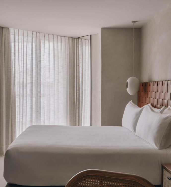 A neatly made bed with white sheets with a leather headboard and curtains over the windows