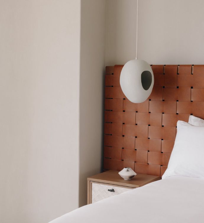 Corner of a white bed with a rust-colored weaved headboard and a white hanging light fixture