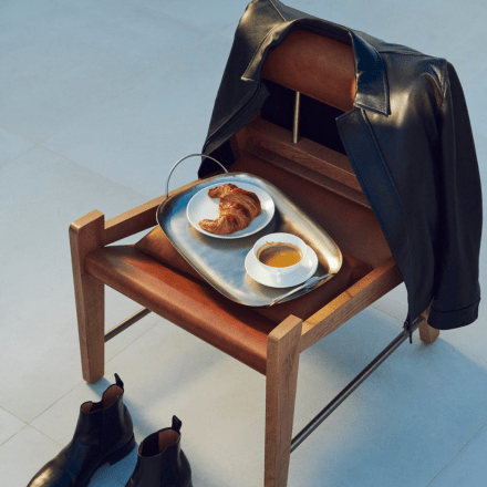 Croissant and coffee on a tray on a wooden chair with leather jacket slung over it and boots on the floor