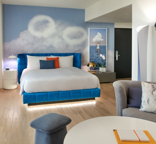 Blue headboard bed with white bedding and a cloud painted wall behind it with white coffee table and grey chair in the foreground