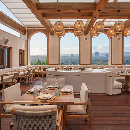 Light wooden and cream dining room with hanging rattan pendant lights, small wooden square table with cream dining chairs in the foreground, and large cream booths in the background with curved windows looking out to Los Angeles cityscape.