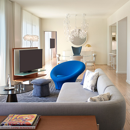 Hotel guest room with a large, grey wraparound couch, blue chair, TV and chandelier.