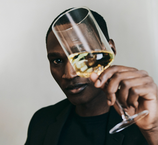 Polished looking man in a black top in front of a grey wall holds a wine glass full of white wine in front of his face and looks into the camera.