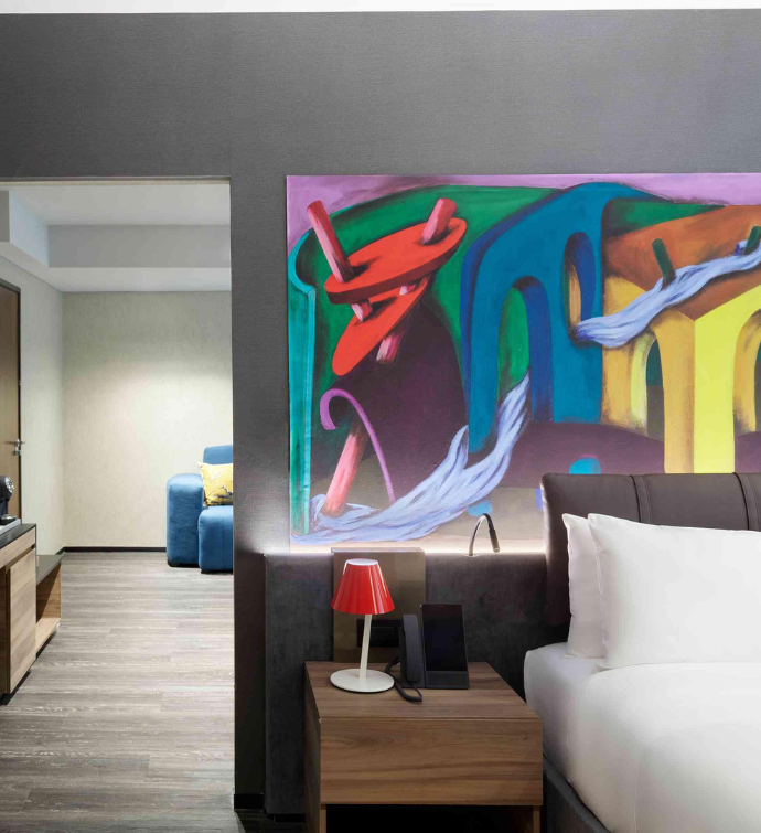 the corner of a bed with bright modern art behind the headboard with a door leading to a separate room