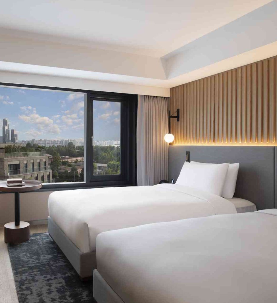 Two white twin beds with grey headboards on a grey patterned rug with a window that overlooks the city with beige curtains