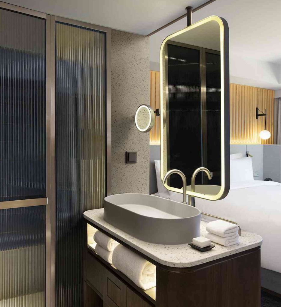 Modern bathroom with a grey sink, marble top, towels folded beneath, a lit up mirror, and a walk-in shower in the corner