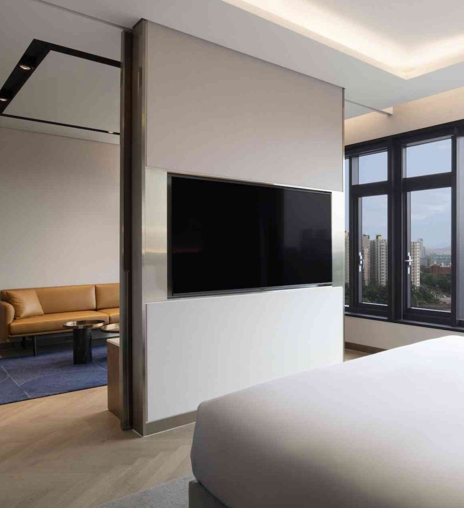 Corner of a white bed facing a cream wall with a tv on it, the room beyond the wall has a tan sofa and coffee table, and windows to the right look out to a city