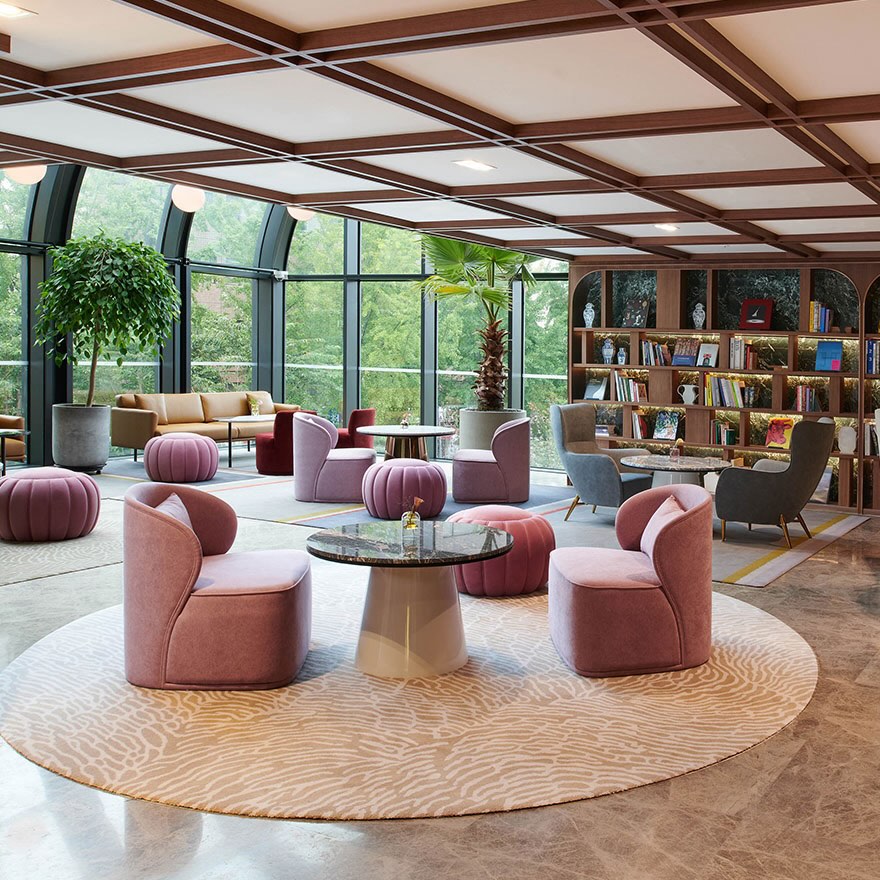 Bright lounge space with pink lounge chairs, grey lounge chairs, and pink ottomans on a beige patterned rug and a marble table