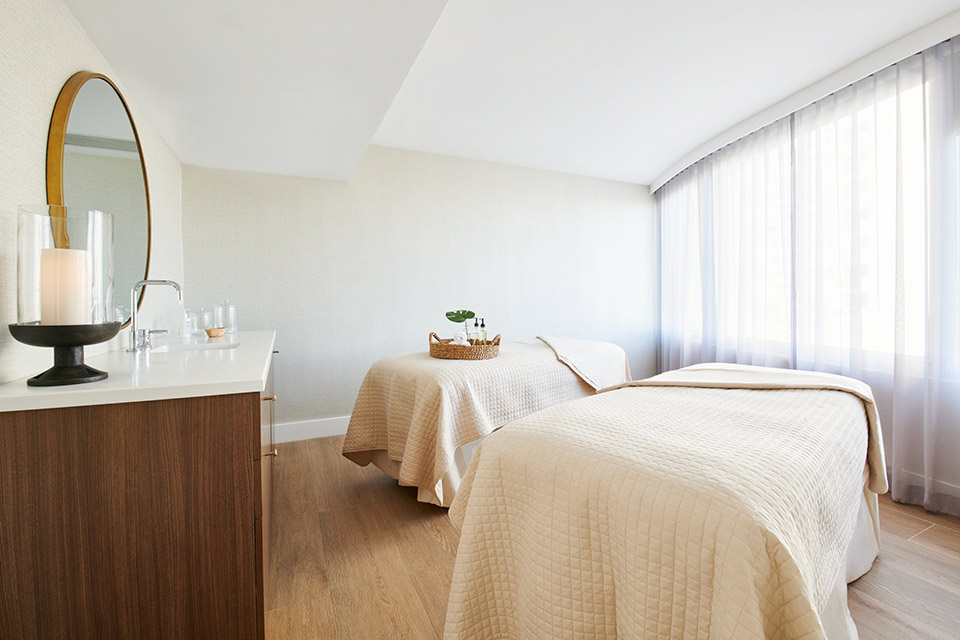 A naturally lit couples massage treatment room with two massage beds and a mirror.