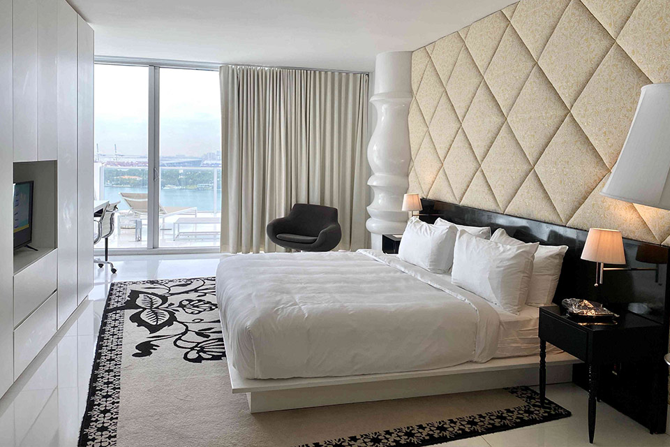 Hotel room at Mondrian South Beach showing a king size bed, a beige quilted pattern on the wall, and floor to ceiling windows with a view of the bay.