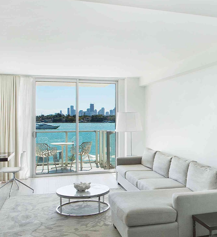 Grey sectional sofa and glass/metal coffee table with flowers on top on a patterned grey rug and a glass sliding door out to a balcony overlooking the water