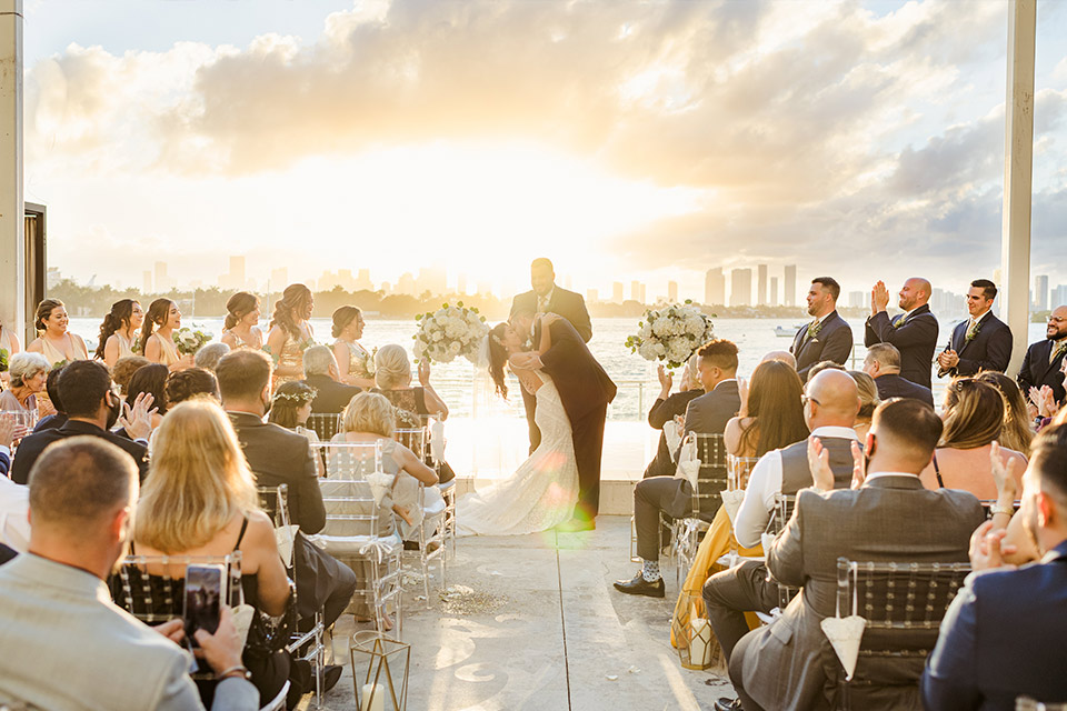 Wedding ceremony overlooking the bay during sunset, with a bride and groom kissing while the bridal party, guests and officiant clap.