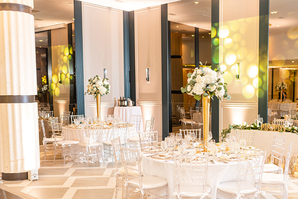 A large room set-up for a wedding with round tables, clear chairs and elegant floral arrangements in tall, gold vases.