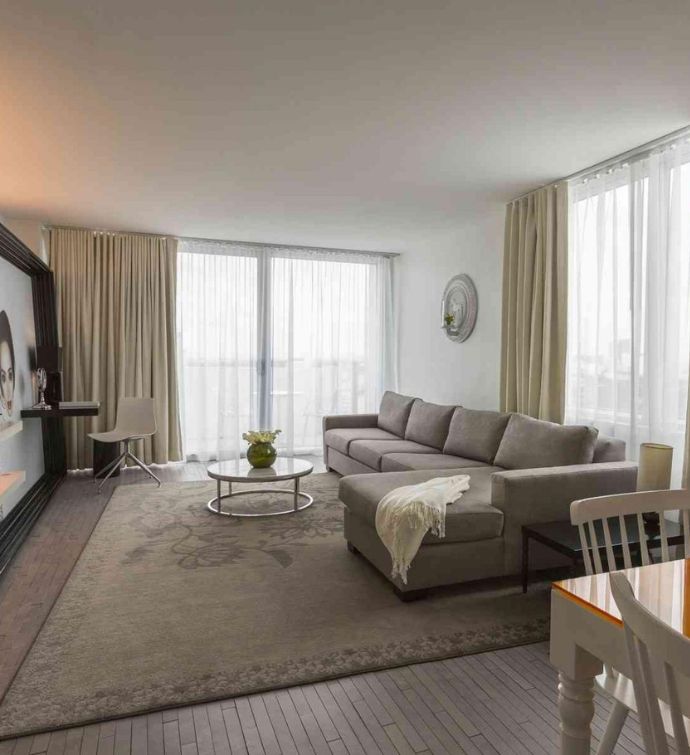 Beige sectional on a beige patterned rug with a coffee table with a plant on it and balcony with curtains in the background