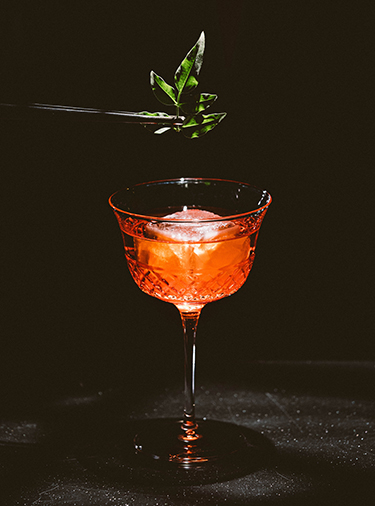 Red cocktail that is being garnished with an herb using a pair of tweezers