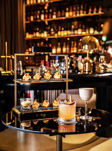 Bites placed on a double-tiered gold and black tray, with two cocktails in front of it and a bar in the background