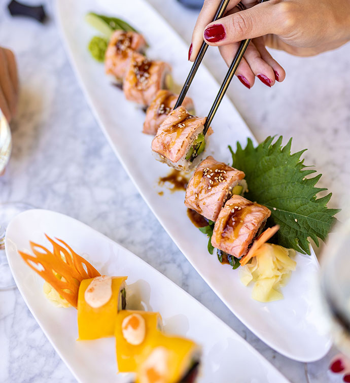 Two platters of sushi rolls from Mr Nakamoto - one topped with mango, one topped with salmon -with 2 pairs of hands picking up a piece with chopsticks