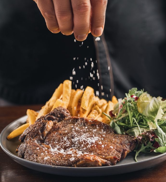 Hand putting salt on a steak and fries with a side salad