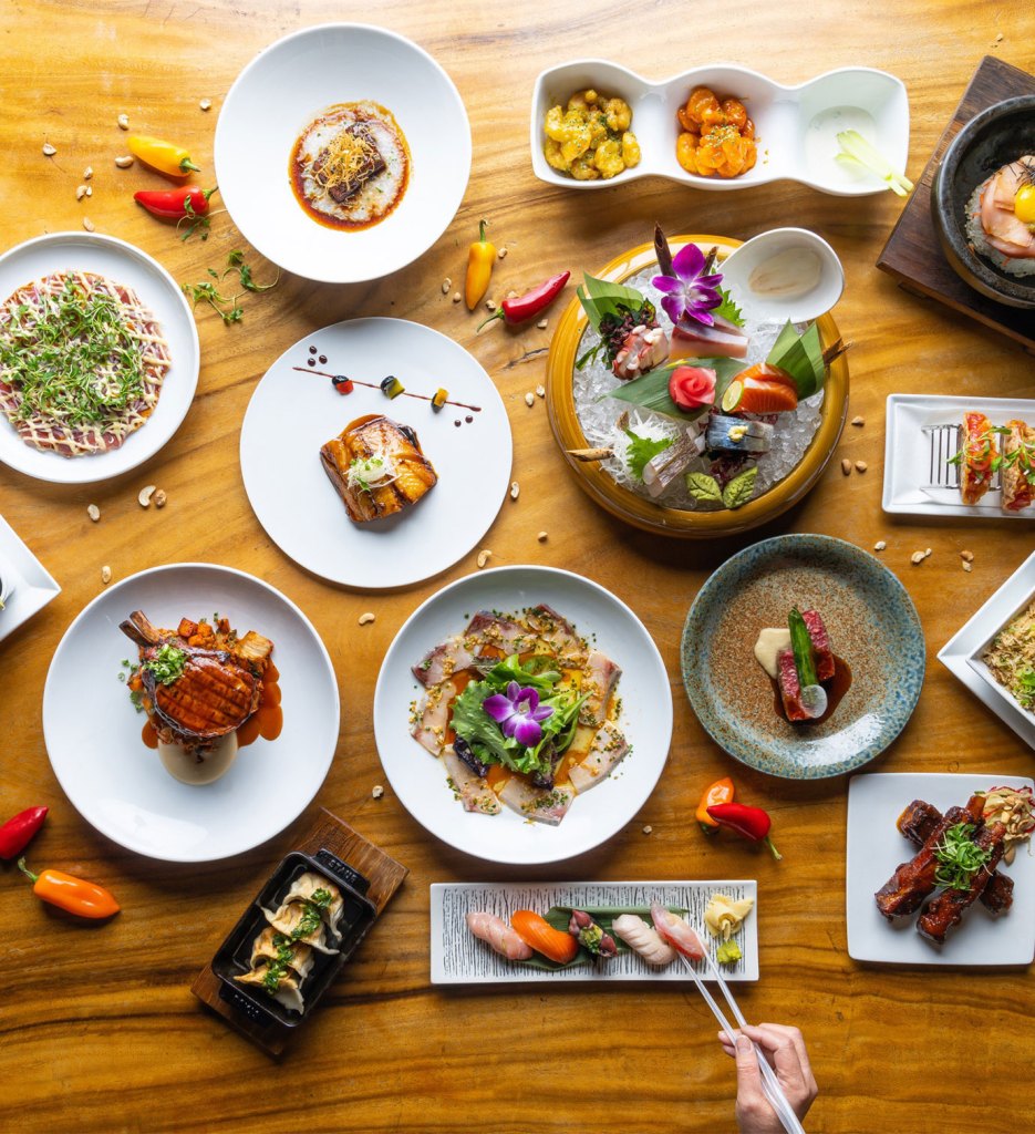 Multiple Japanese dishes on different shaped and colored plates sit on a light wooden tabletop