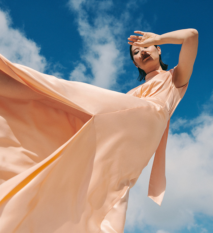 Woman in long peach dress with hand over her face and blue sky with clouds in the background