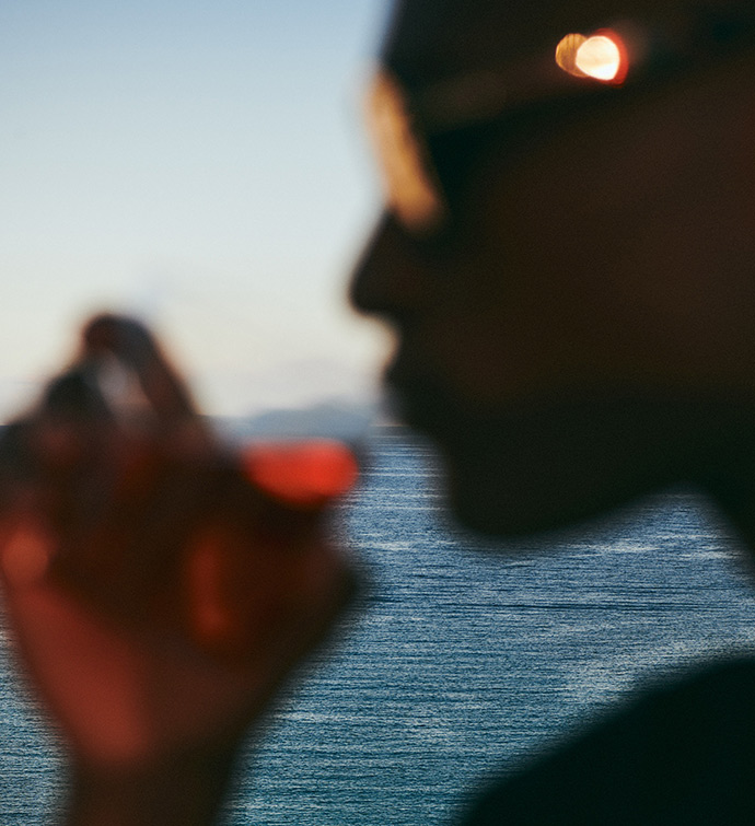 Close up image of person putting a cocktail up to their mouth with an ocean in the background