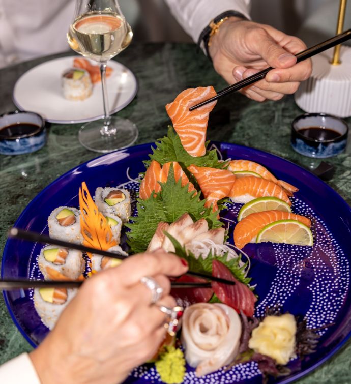 Sushi and sashimi on a blue plate with a glass of wine in the background and two hands with chopsticks picking up sushi