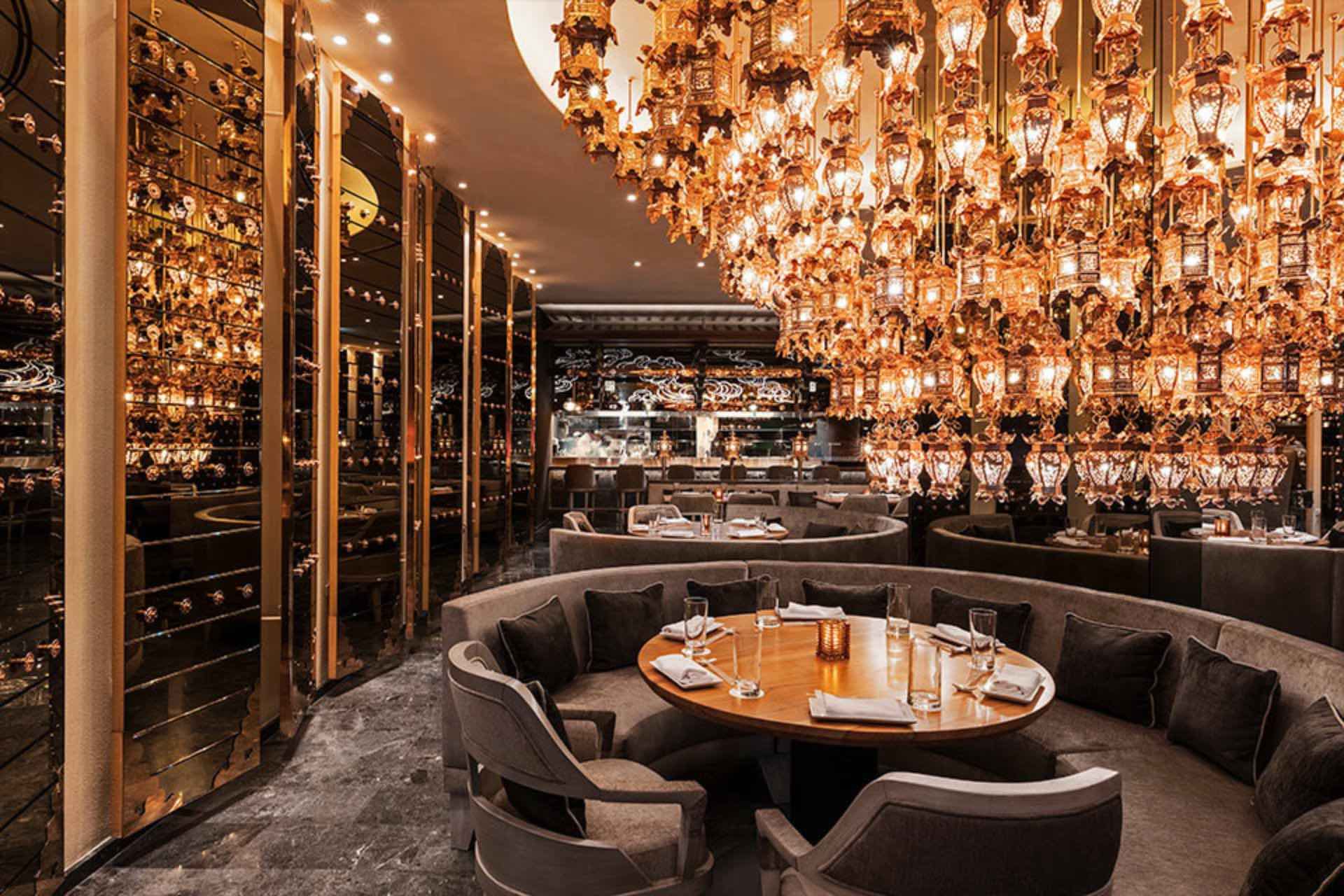 Dining room with large, elaborate gold lights hanging in rows, a grey rounded dining booth, circular wooden table, and two grey dining chairs with other similar tables in the background.