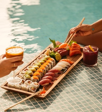 Large plate of sushi and two cocktails with hands in the corner of the image and a pool in the background