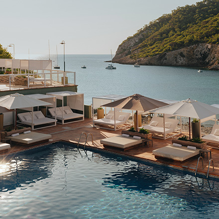 Blue pool with large cream-colored daybeds and cream umbrellas on a pool deck with blue sea and tree-covered mountains in the background.