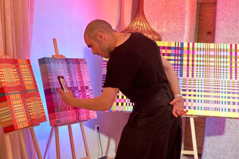 Man in black shirt and pants looking at paintings on easels and taking a photo on his iPhone