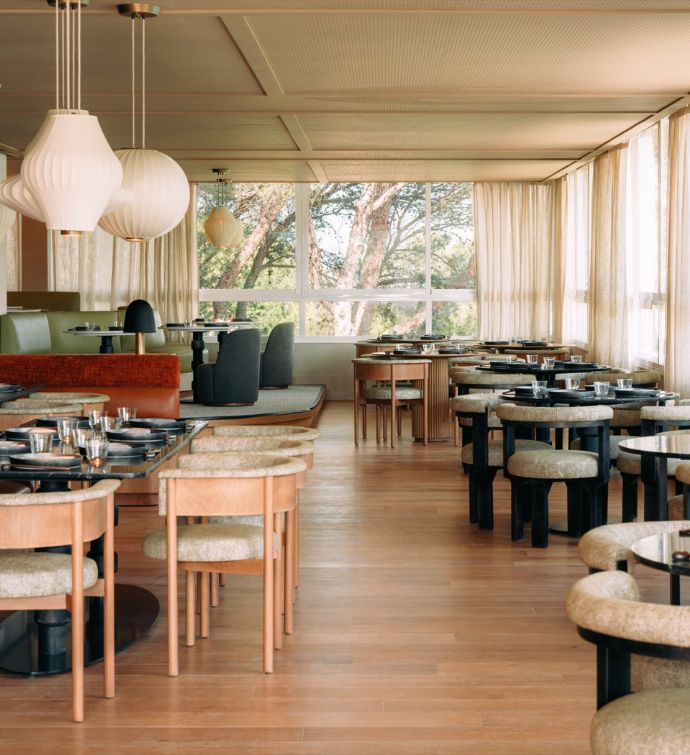 Large bright dining room with various colors and shapes of dining tables and dining seating with cream hanging lanterns and a window in the back looking out to trees