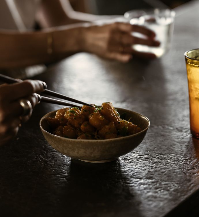 Bowl of tempura with chopsticks digging into it on a dark table with hands holding a glass in the background