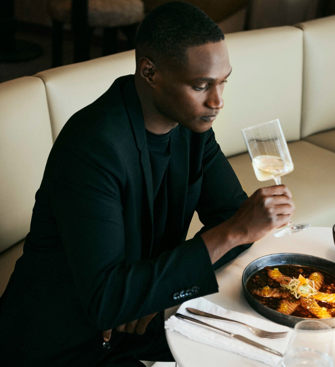 a man sittiing on a tan leather chair at a table looking at his wine glass with fresh food plated in front of him