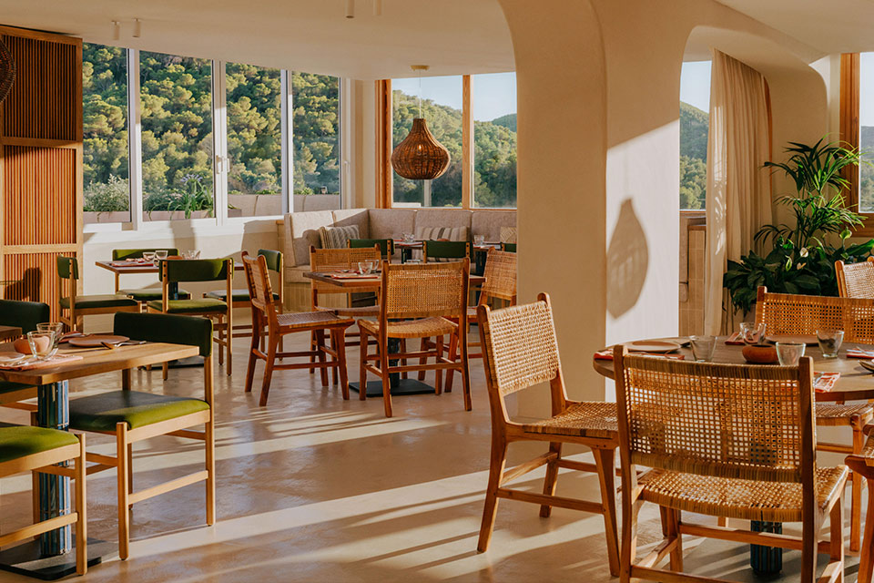 Large dining room dripping in natural sunlight with small wooden tables and wicker chairs spread throughout.